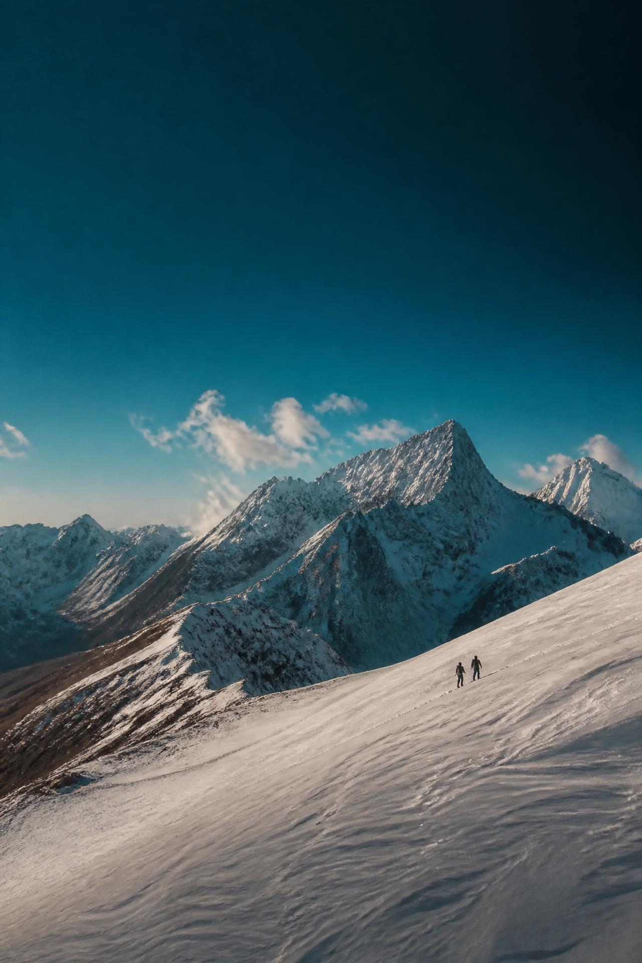 Silhuetas caminhando na neve sob céu azul intenso, com montanhas ao fundo
